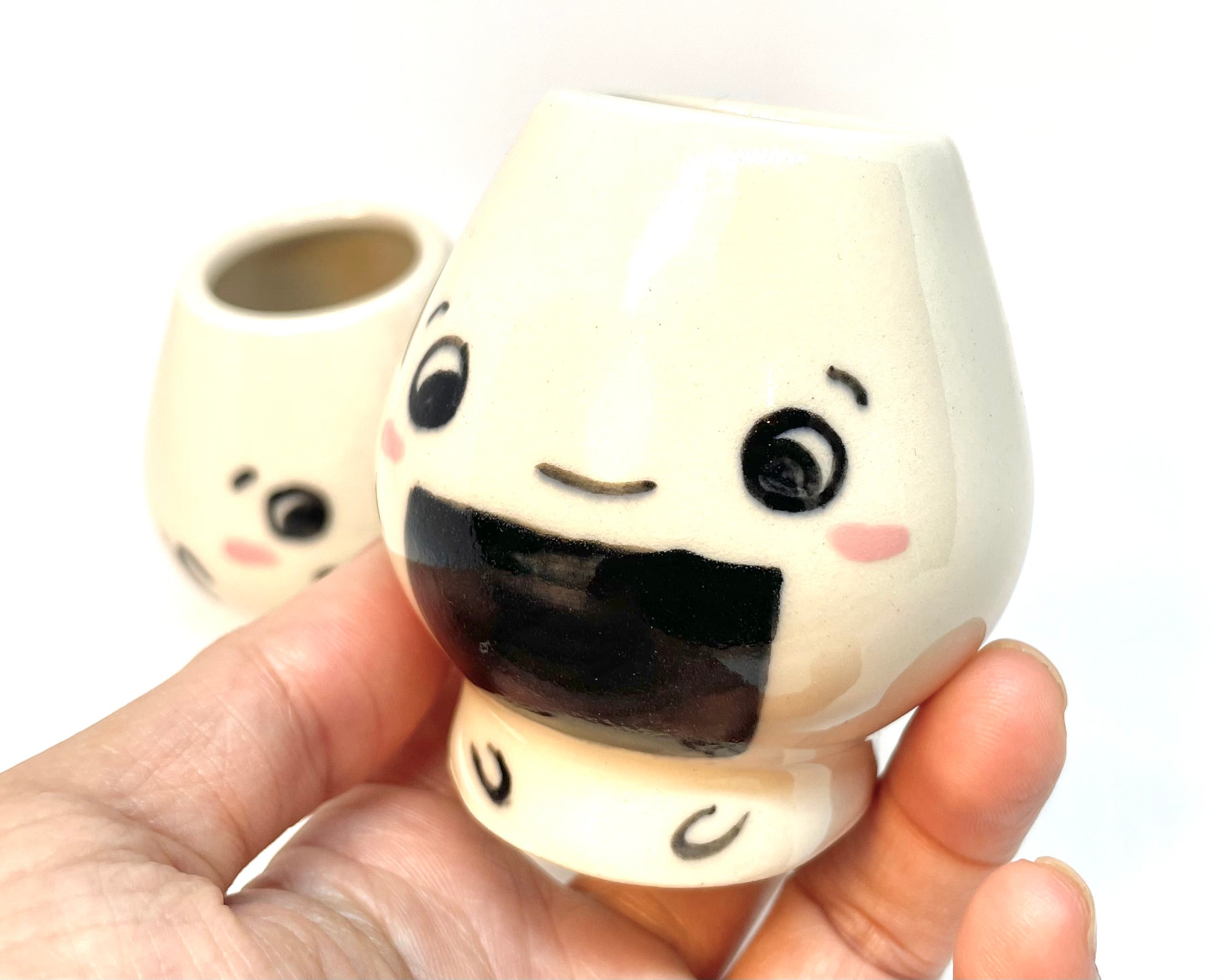 Two small ceramic matcha whisk holder with onigiri faces held by a hand against a white background
